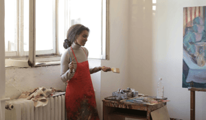 WOman in painting apron standing in front of window with paing on pallet and holding paint brush
